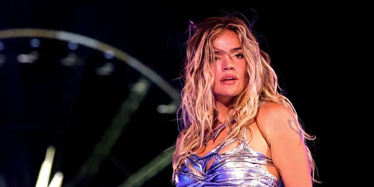 Karol G, con su melena rubia al viento, brilla en el escenario. Su atuendo plateado y el ambiente nocturno sugieren un concierto inolvidable de su gira.