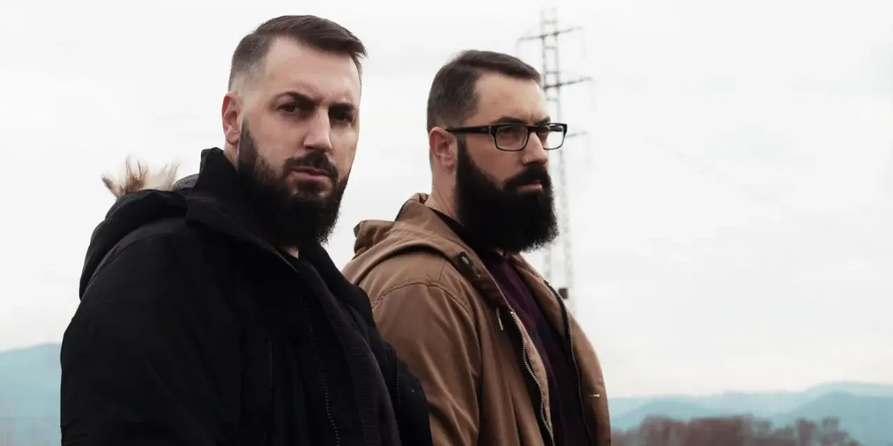 Dos hombres con barba posan con seriedad. Uno viste chaqueta negra, el otro marrón. Parecen listos para un rap de "falsa alarma".