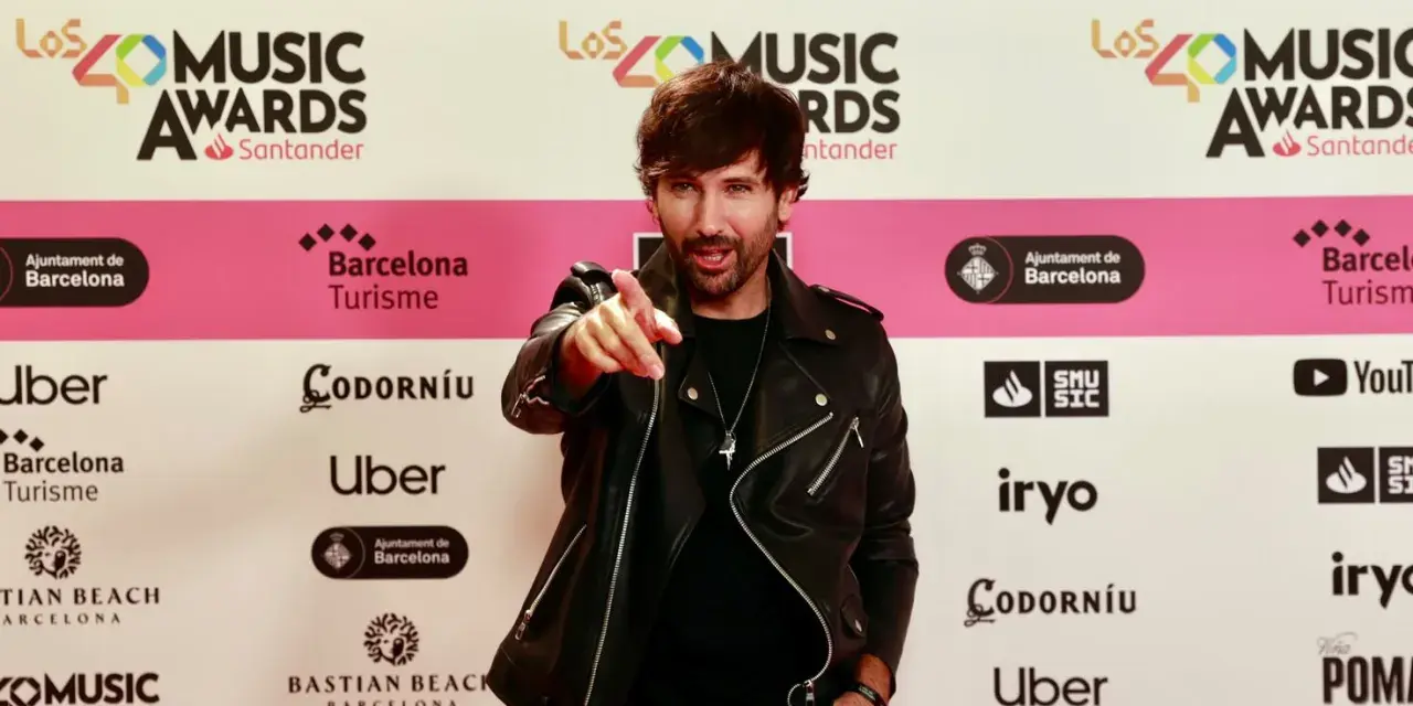 Hombre con chaqueta de cuero negro señala a la cámara en una alfombra roja de Los40 Music Awards. ¡Parece que ha pescado el premio!