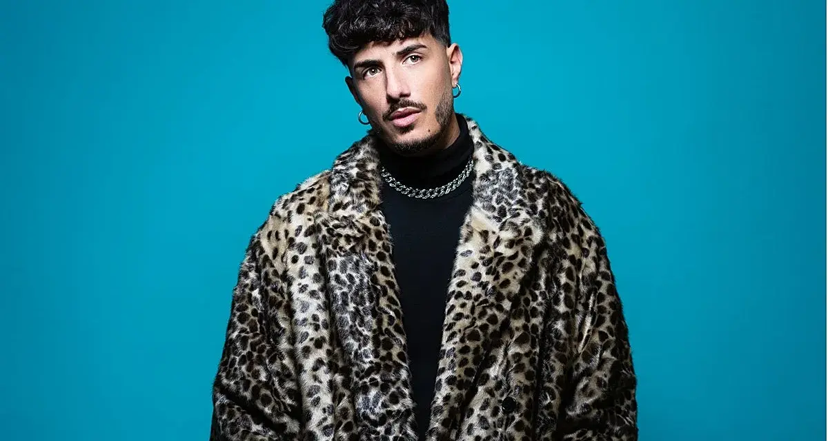 Wade DJ, con su abrigo de leopardo y cadena plateada, posa con confianza ante un fondo azul vibrante.