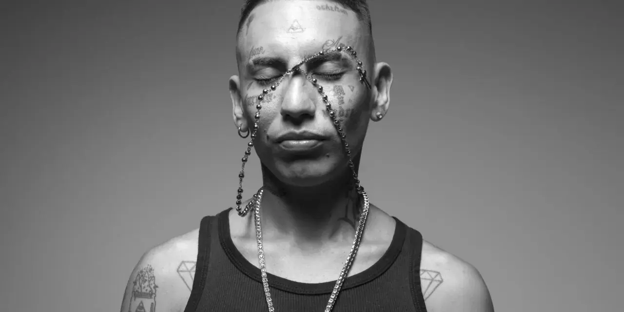 Un joven con tatuajes en la cara y el cuerpo, luciendo un collar de cuentas y una cadena de diamantes. Su estilo neo pistea barcelona es inconfundible.