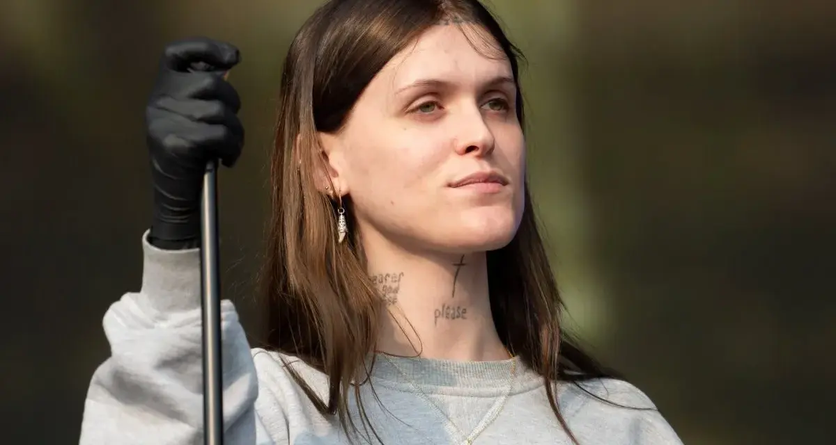 Ethel Cain, con guantes negros y un collar de cruz, mira hacia un lado con una expresión pensativa.