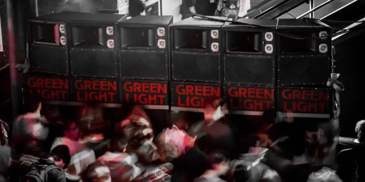 Altavoces apilados con luces rojas "GREEN LIGHT" y gente borrosa bailando. Un sistema de sonido que parece dar problemas.