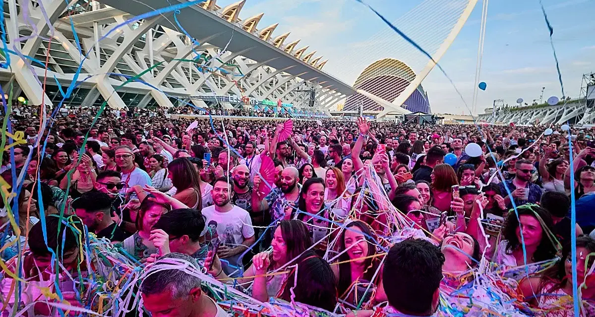 Multitud disfrutando de un fan music fest bajo confeti y luces, con arquitectura moderna de fondo.