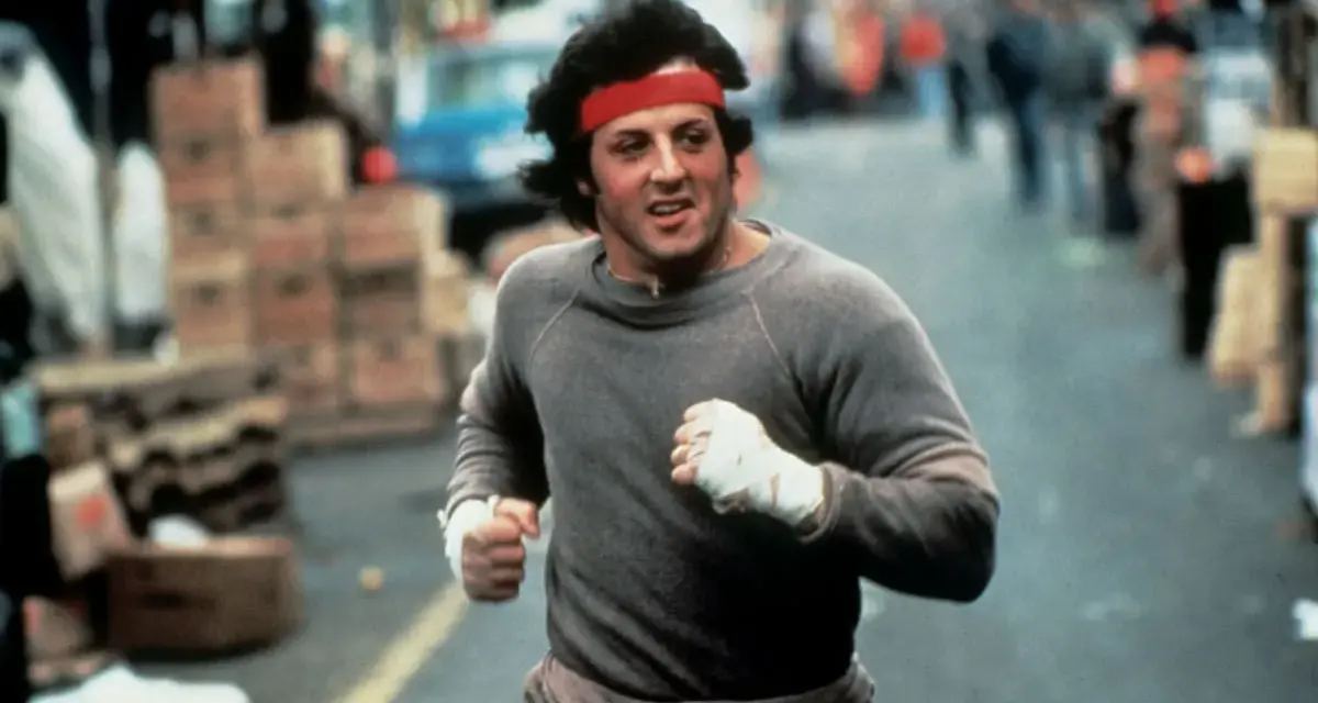 Rocky Balboa corre por la calle, con su icónica banda roja en la cabeza. La banda sonora de Rocky resuena en tu mente.
