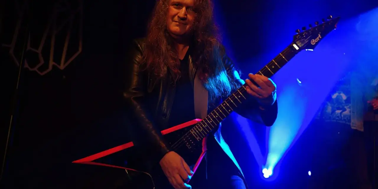 Guitarrista con pelo largo y chaqueta de cuero, tocando una guitarra eléctrica negra con detalles rojos. El escenario está iluminado con luces azules, creando un ambiente de rock.