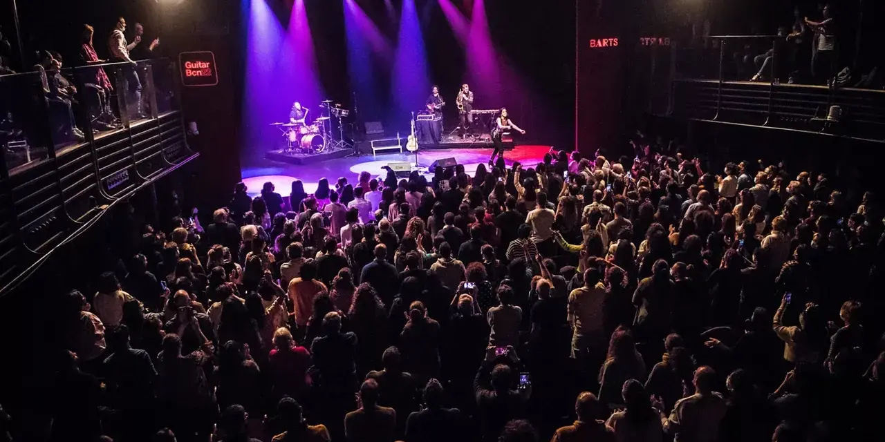 Un concierto en Guitar BCN ilumina el escenario con luces púrpuras mientras una multitud entusiasta graba el momento.