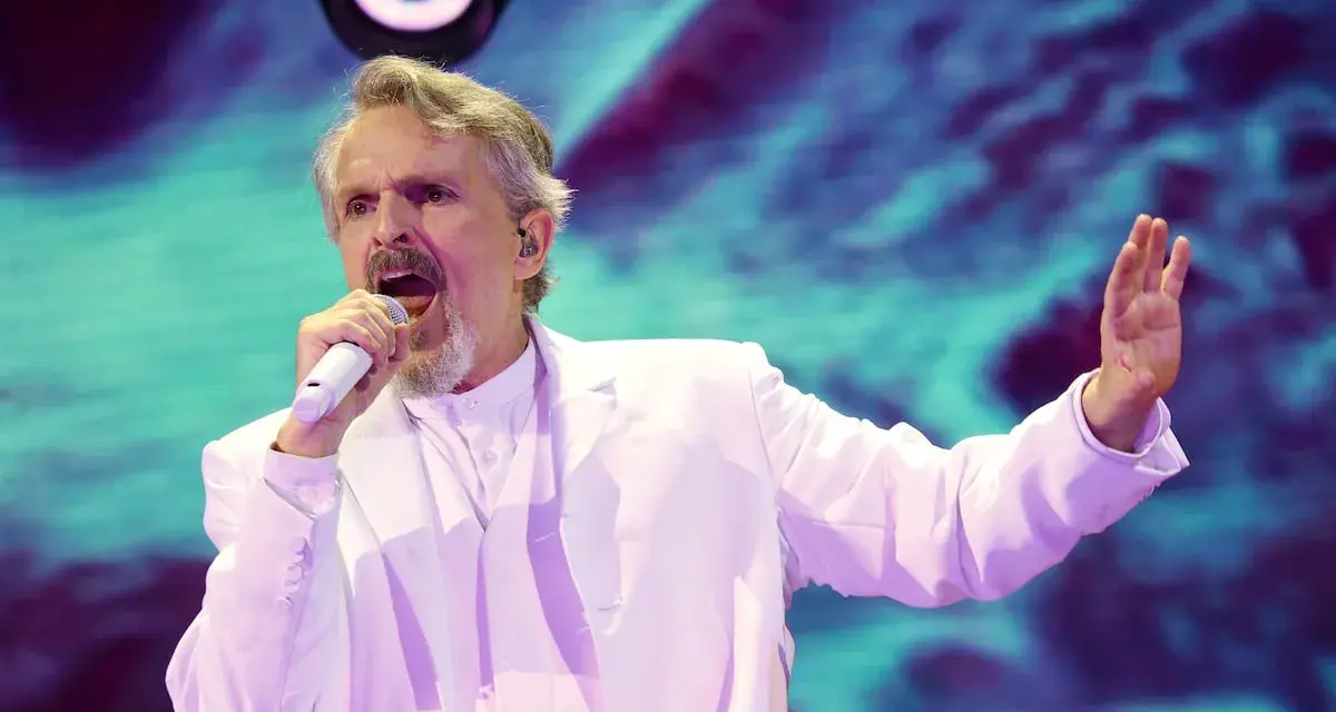 Un hombre con traje blanco canta en un escenario con luces vibrantes. ¡Murcia on!