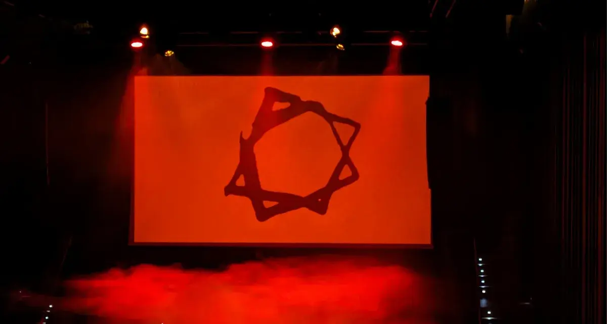 Símbolo estilizado en pantalla naranja, con humo rojo y luces de escenario. Podría ser parte de una dellafuente biografia visual.