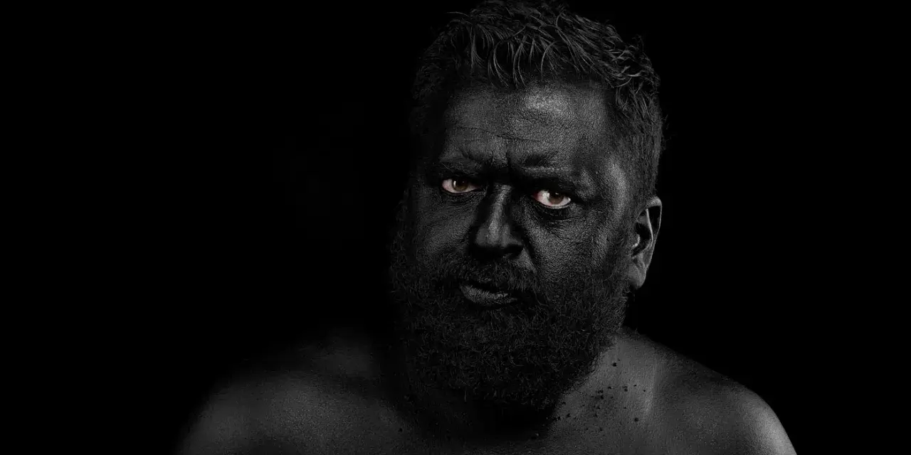 Hombre con el rostro y cuerpo cubiertos de pintura negra, ojos penetrantes. Parece un guerrero ancestral, don fluor.