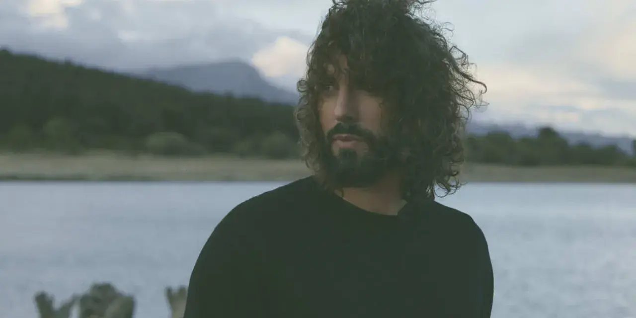 Un hombre con cabello rizado y barba mira hacia el horizonte, entre el miedo y el paraíso de un paisaje acuático y montañoso.