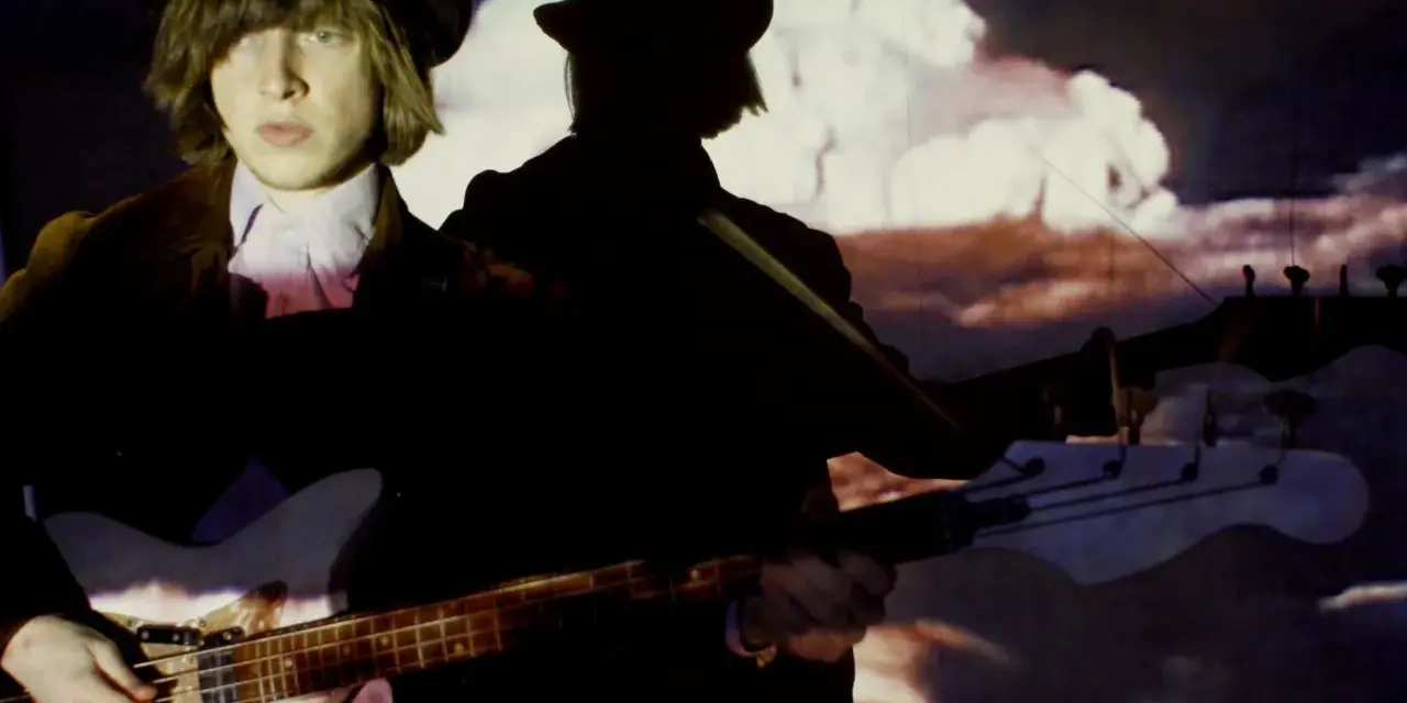 Jacco Gardner, con sombrero y chaqueta, toca el bajo frente a una proyección psicodélica.