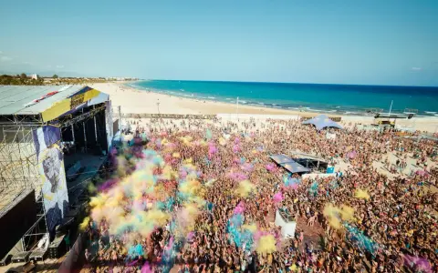 Multitud vibrante en el Arenal Sound Valencia, lanzando polvo de colores bajo el sol. El mar azul y la playa de fondo crean un escenario festivo.