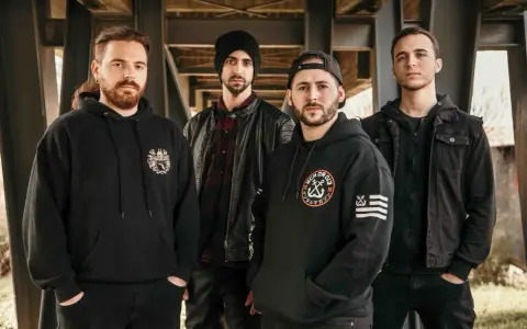 Cuatro hombres con sudaderas y chaquetas negras posan bajo una estructura metálica. Sus miradas intensas parecen **blaze out** con determinación.