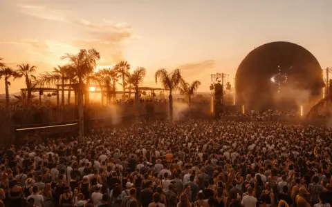 Multitud en el festival Sophie, disfrutando del atardecer con palmeras y una gran esfera como escenario.