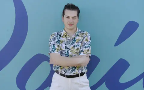 Joven con camisa tropical y pantalones claros, brazos cruzados, frente a un mural azul con formas orgánicas. Parece un miembro del ortiga grupo.