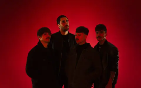 Cuatro hombres posan ante un fondo rojo vibrante. Parecen una banda, quizás "los flamingos", con sus miradas intensas y atuendos modernos.