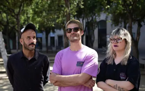 Miembros de Els Catarres posan al aire libre. El hombre de la izquierda lleva gorra y camisa negra. El del centro, gafas de sol y camiseta lila. La mujer de la derecha, gafas y camiseta negra.