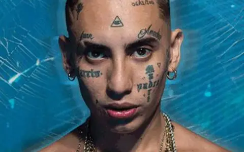 Joven con tatuajes en la cara, incluyendo un ojo dentro de un triángulo, y cadenas de oro. Su estilo neo pistea madrid es inconfundible.