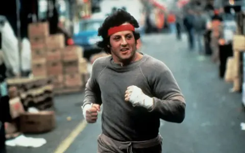 Rocky Balboa corre por la calle, con su icónica banda roja en la cabeza. La banda sonora de Rocky resuena en tu mente.