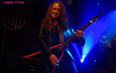 Guitarrista con pelo largo y chaqueta de cuero, tocando una guitarra eléctrica negra con detalles rojos. El escenario está iluminado con luces azules, creando un ambiente de rock.