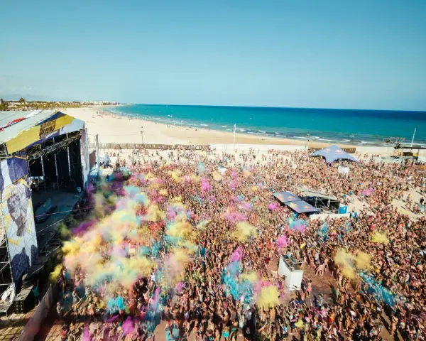 Multitud vibrante en el Arenal Sound Valencia, lanzando polvo de colores bajo el sol. El mar azul y la playa de fondo crean un escenario festivo.