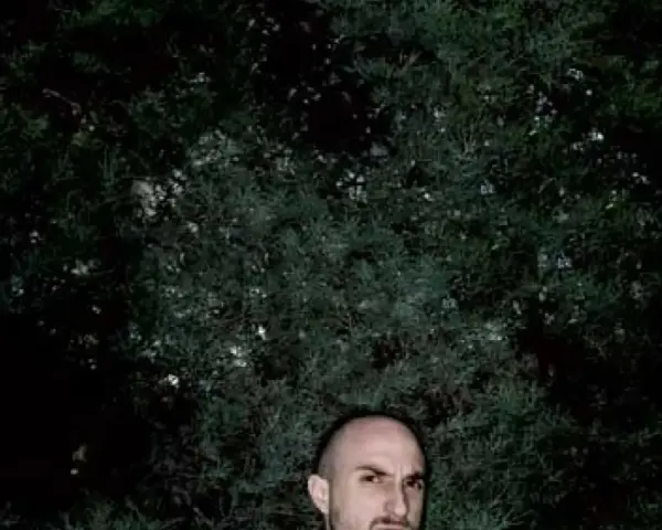 Un hombre calvo, con mirada intensa, posa frente a un fondo de follaje oscuro. Podría ser un sicario rapero, listo para soltar rimas letales.