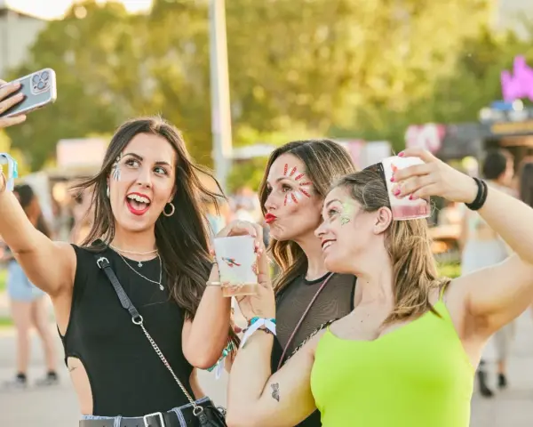 Tres amigas con maquillaje festivo y bebidas en un festival de verano.