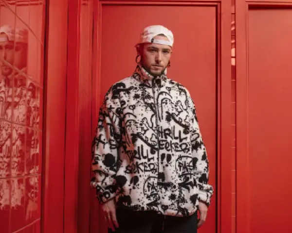 El cantante Kaze, con gorra blanca y chaqueta polar con grafitis, posa en un pasillo rojo vibrante.