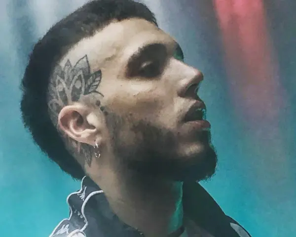 Un joven **hard cantante** con tatuaje en la cabeza y barba, mirando hacia arriba con luces de neón de fondo.