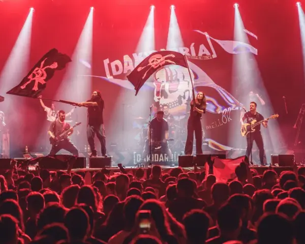 Una banda toca en un escenario iluminado con luces rojas, ondeando banderas piratas. El público, con trece roeis, anima la actuación.