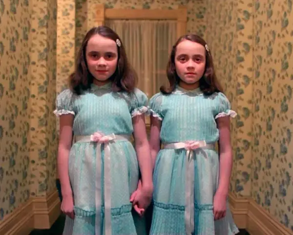 Dos niñas idénticas, vestidas con trajes azules, se toman de la mano en un pasillo con papel tapiz floral. Son las gemelas de "Bso El Resplandor".