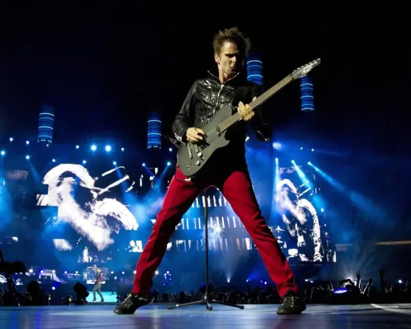Matt Bellamy de Muse España, con pantalones rojos y chaqueta negra, toca la guitarra en un escenario iluminado por luces azules y pantallas gigantes.