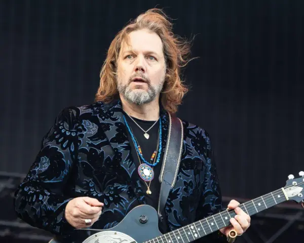 Rich Robinson, guitarrista con cabello largo y barba, luce un saco negro con estampado floral azul y varias cadenas.
