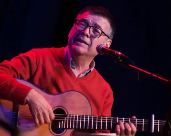 Mario Díaz, con gafas y un suéter rojo, toca la guitarra y canta frente a un micrófono en un escenario con luces tenues.