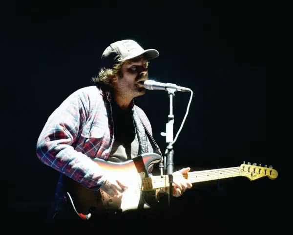 Neil Halstead, con gorra y camisa a cuadros, toca la guitarra y canta en un concierto.