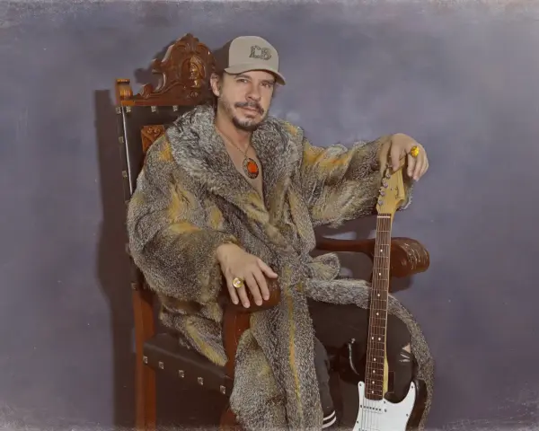 Luis Brea, con gorra y abrigo de piel, sentado en un trono con una guitarra.