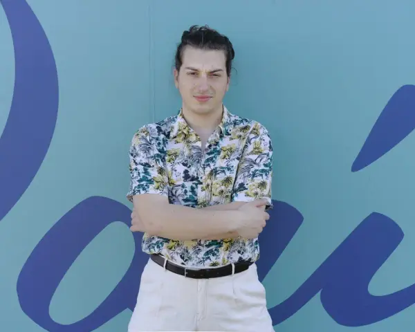 Joven con camisa tropical y pantalones claros, brazos cruzados, frente a un mural azul con formas orgánicas. Parece un miembro del ortiga grupo.