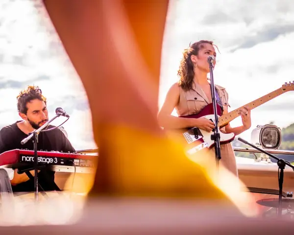 Maria y Feu, una banda en vivo, tocan en un escenario al aire libre. Ella canta y toca la guitarra, él en los teclados.