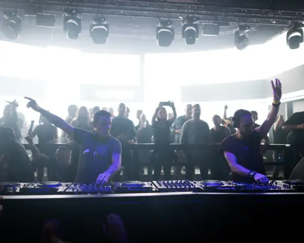 Dos DJs actuando en un club con público al fondo.