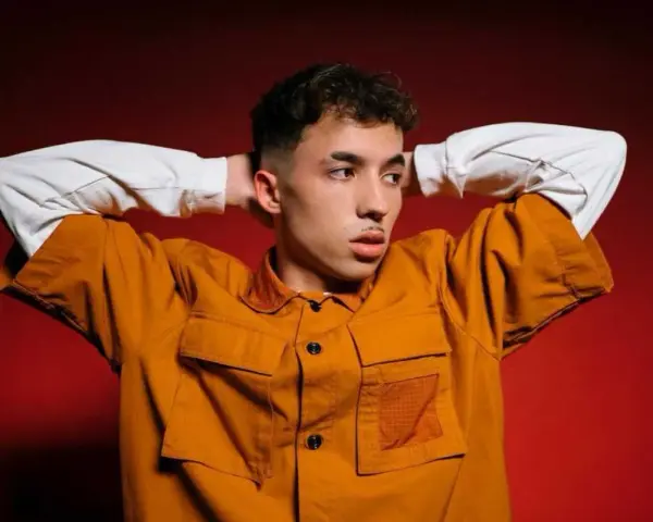 Joven con las manos detrás de la cabeza, vistiendo una chaqueta naranja y una camiseta blanca. Parece un pole cantante listo para actuar.