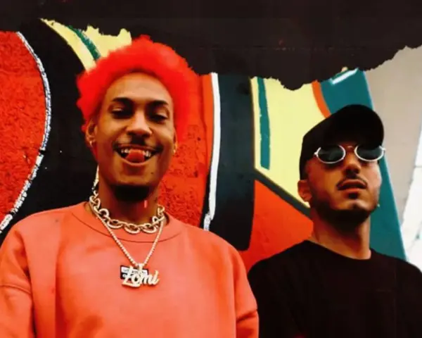 Dj Ballesteros y su colega posan frente a un mural vibrante. El primero, con cabello rojo y un collar dorado, sonríe con la lengua afuera.