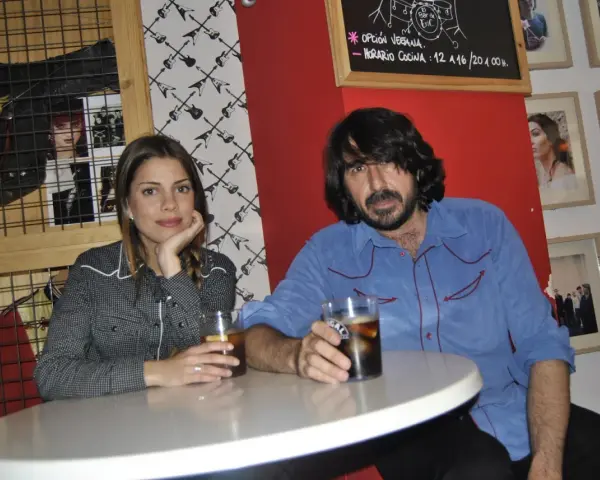Pareja en un bar con decoración rockera. Ella, con camisa de cuadros, apoya la barbilla en la mano. Él, con camisa azul, sostiene un vaso. La furia trinidad de la música inspira el ambiente.