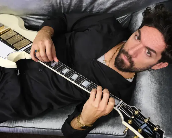 El cantante Siloe, con barba y cabello oscuro, toca una guitarra eléctrica blanca con detalles dorados, recostado en un sofá de terciopelo gris.