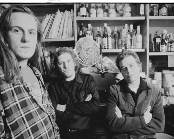 Tres hombres posan en blanco y negro, con estanterías de discos y botellas detrás.