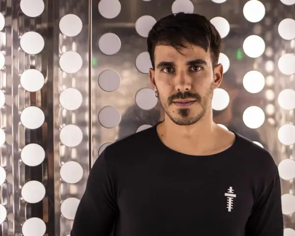 Raúl Pacheco, DJ, posa con una camiseta negra frente a un fondo metálico con círculos blancos brillantes.
