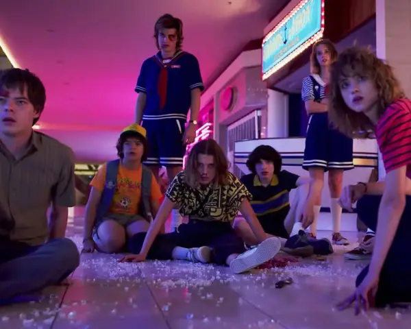 Los personajes de Stranger Things, con Eleven al frente, sentados en el suelo de un centro comercial, evocando las icónicas canciones de la serie.