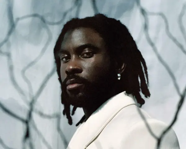 Retrato de un rapero español con rastas y un pendiente plateado, vistiendo una chaqueta clara.
