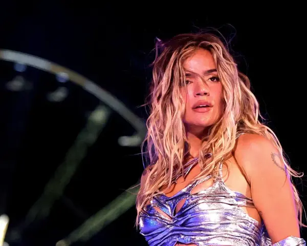 Karol G, con su melena rubia al viento, brilla en el escenario. Su atuendo plateado y el ambiente nocturno sugieren un concierto inolvidable de su gira.