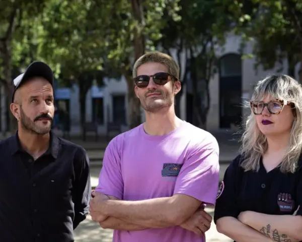 Miembros de Els Catarres posan al aire libre. El hombre de la izquierda lleva gorra y camisa negra. El del centro, gafas de sol y camiseta lila. La mujer de la derecha, gafas y camiseta negra.